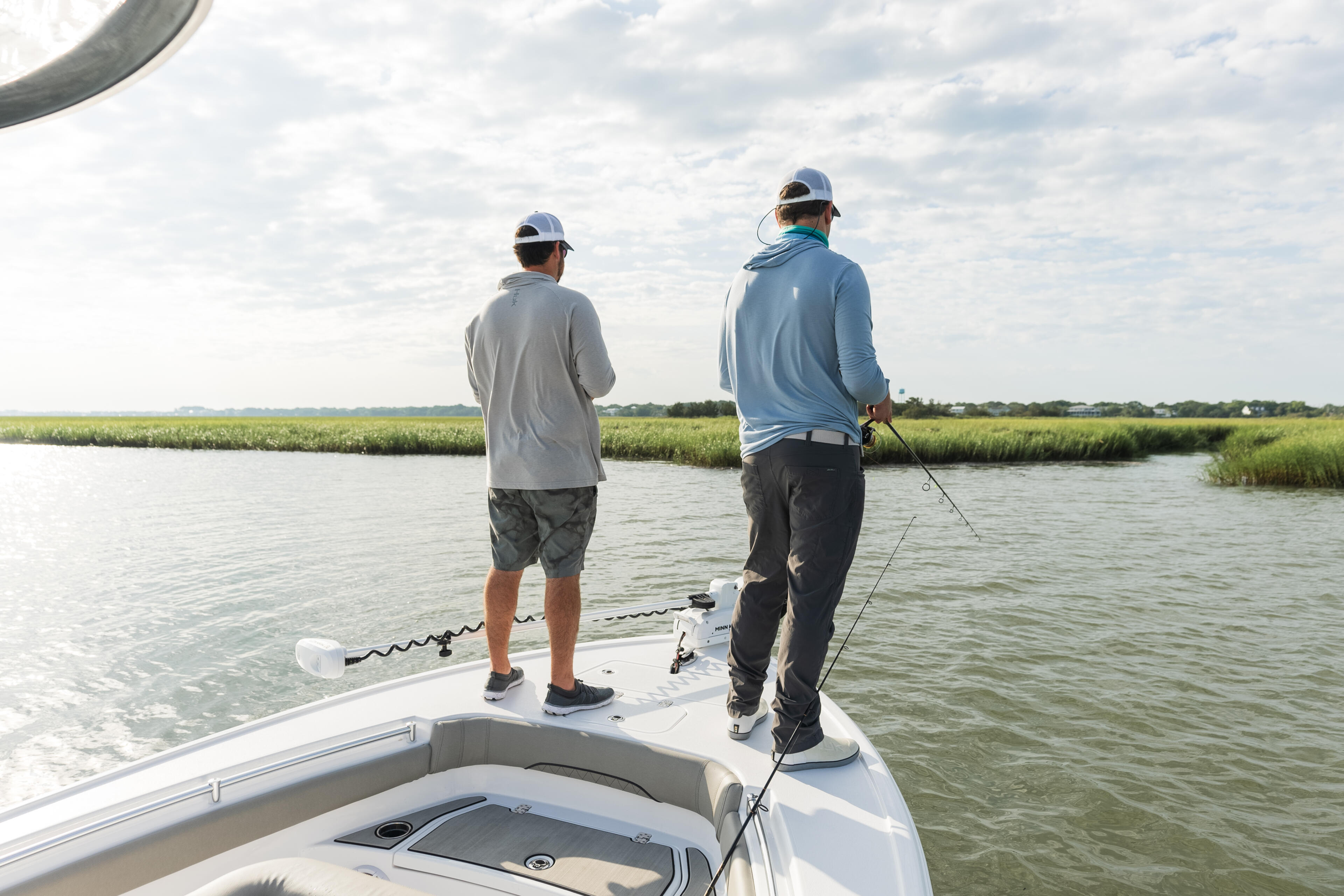 Two anglers standing on the bow of a center console boat in shallow coastal waters with marsh grass visible