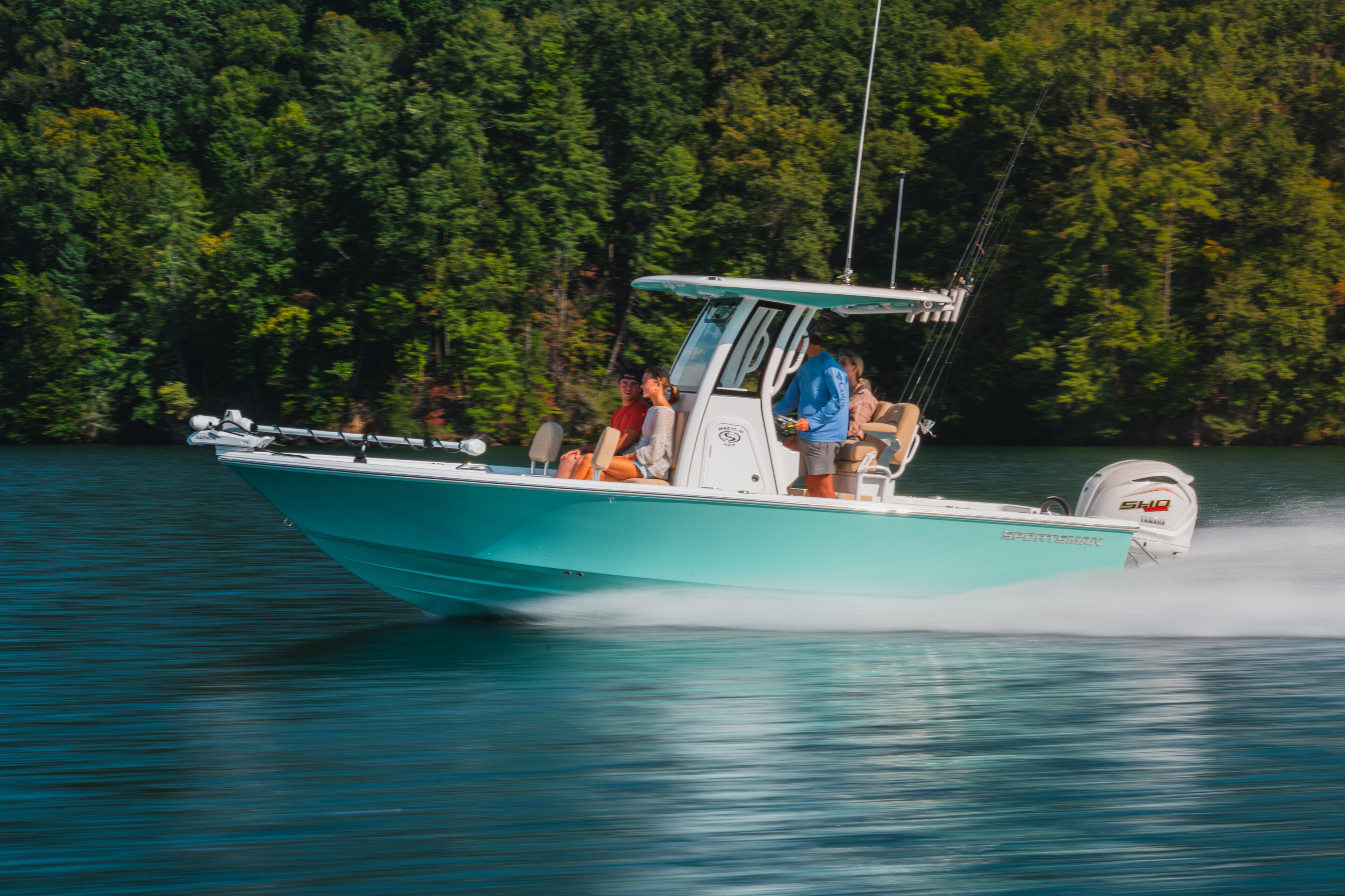 Turquoise fishing boat with passengers cruising at speed past forested shoreline on clear water