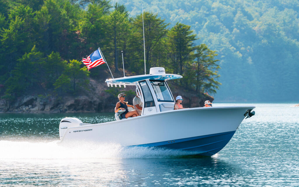 White and blue center console boat running on clear lake water with American flag, forested shoreline in background