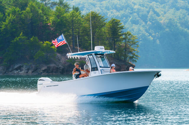 White and blue center console boat running on clear lake water with American flag, forested shoreline in background