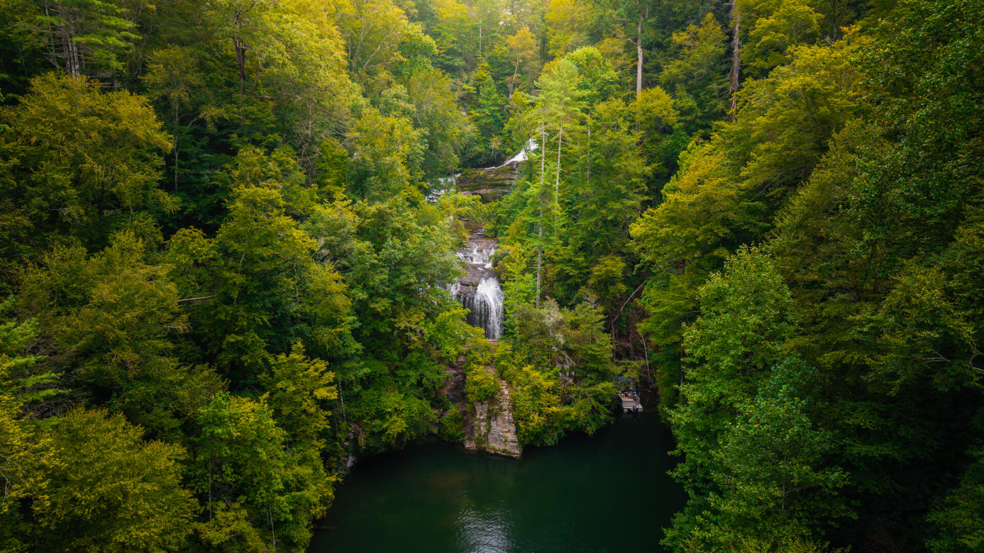Aerial view of a cascading waterfall hidden within dense green forest surrounding a pristine dark water lake