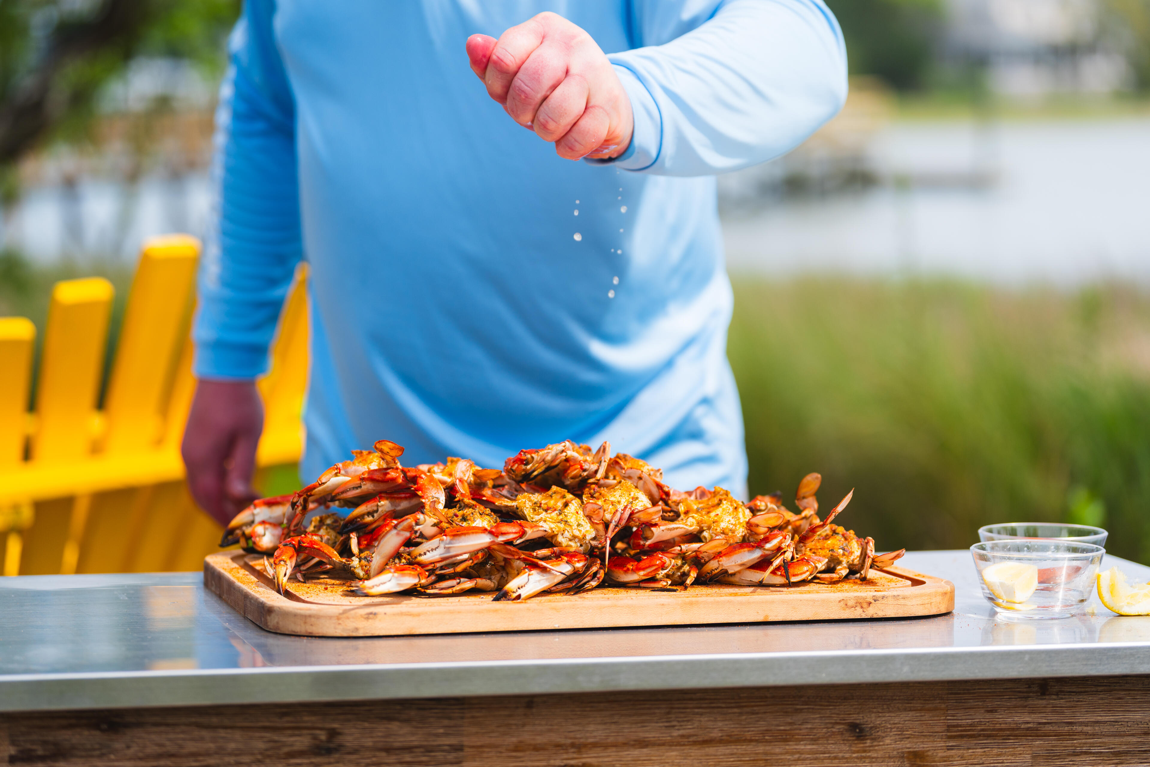 Child in blue shirt reaching toward a pile of cooked crabs on a wooden board at an outdoor waterside table