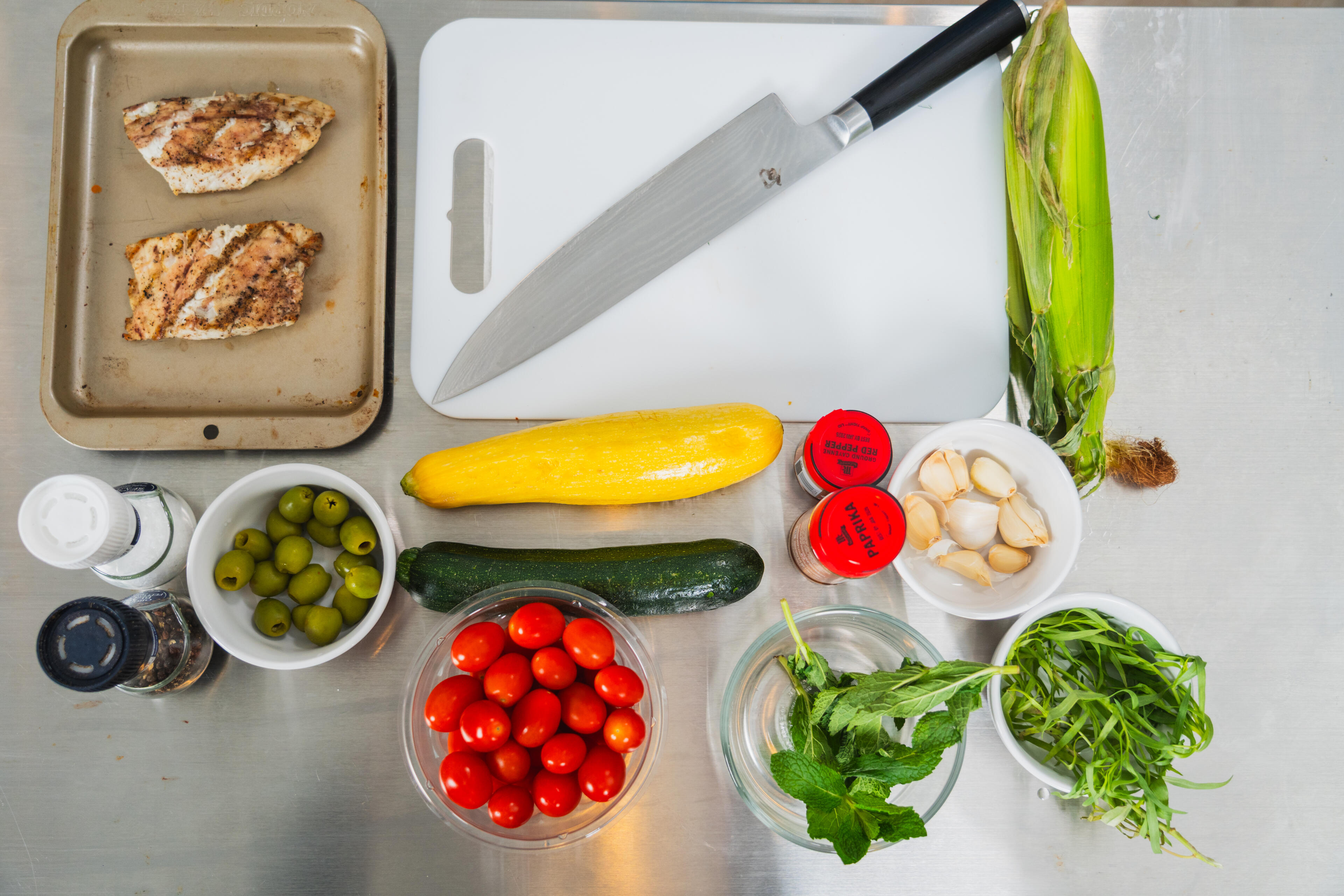 Cooked redfish fillets on a baking sheet surrounded by fresh ingredients including tomatoes, cucumber, corn, olives, garlic, and herbs on a prep counter