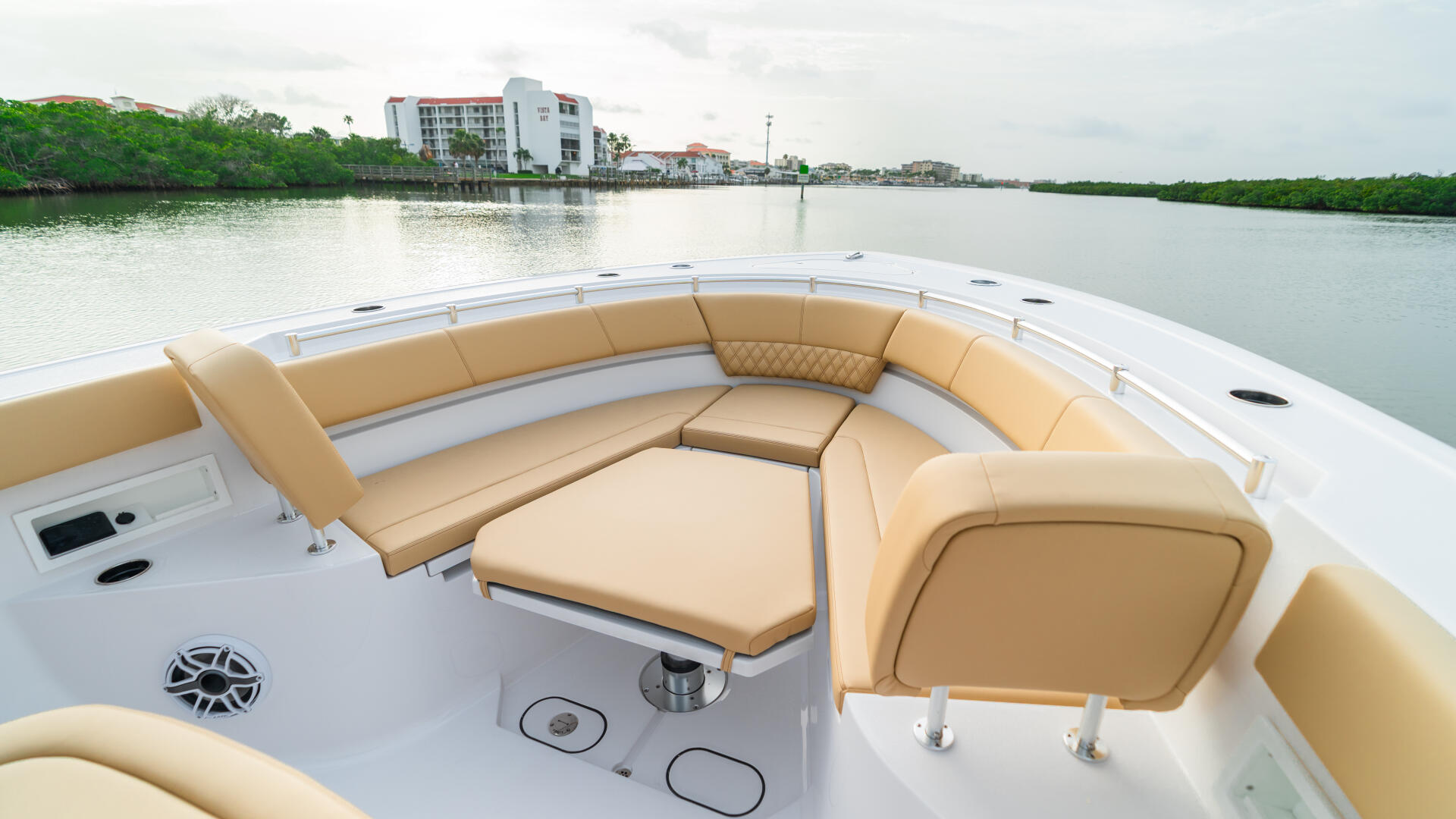 Spacious curved seating area with tan cushions and white helm console overlooking calm waterway and coastal buildings