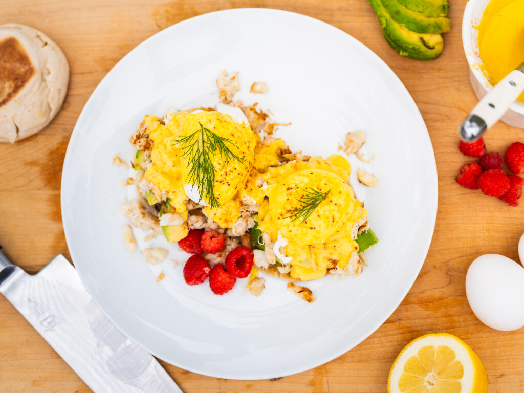 Crab benedict with poached eggs, hollandaise, fresh raspberries and dill on white plate surrounded by ingredients
