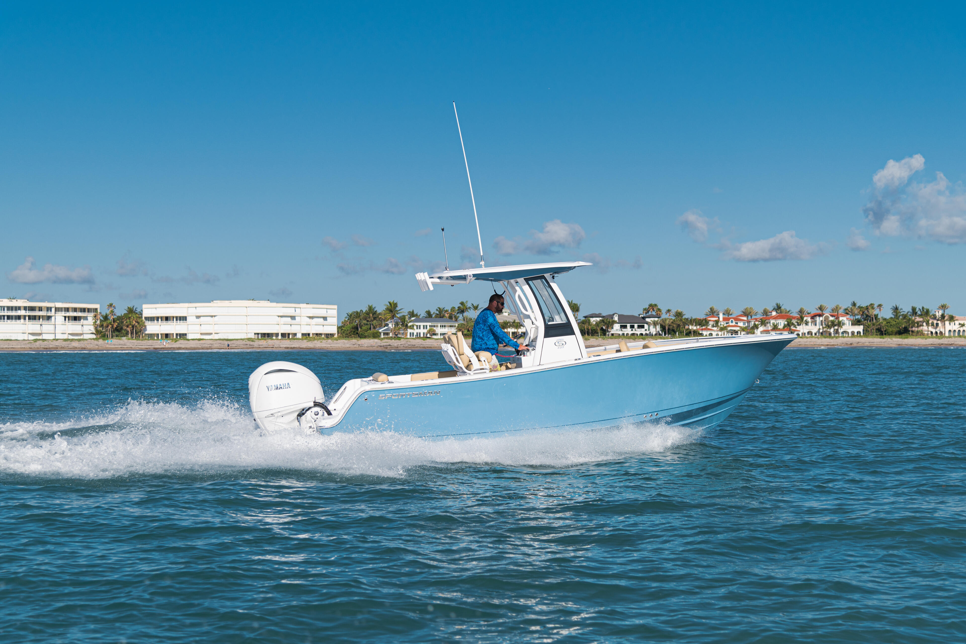 Blue and white center console boat running offshore with hard-top and operator at helm, coastal homes visible on beach in background