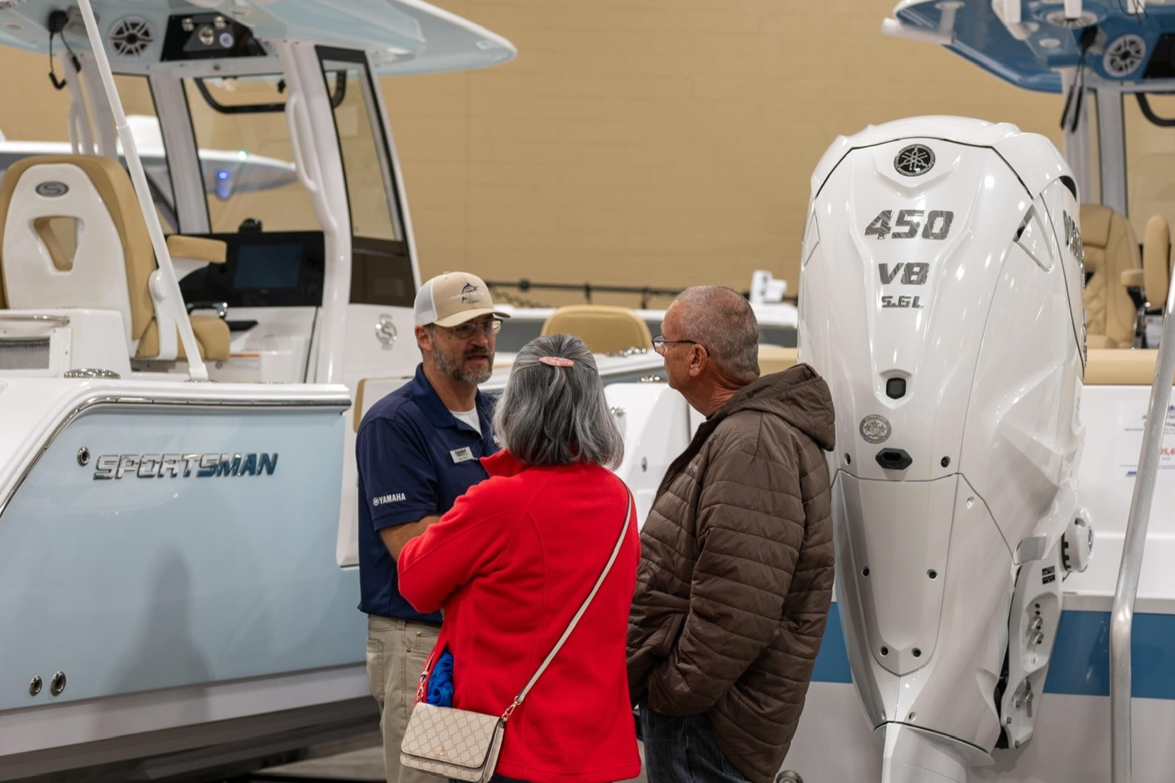 Salesman demonstrates boat features to couple at indoor marina showroom with multiple vessels on display