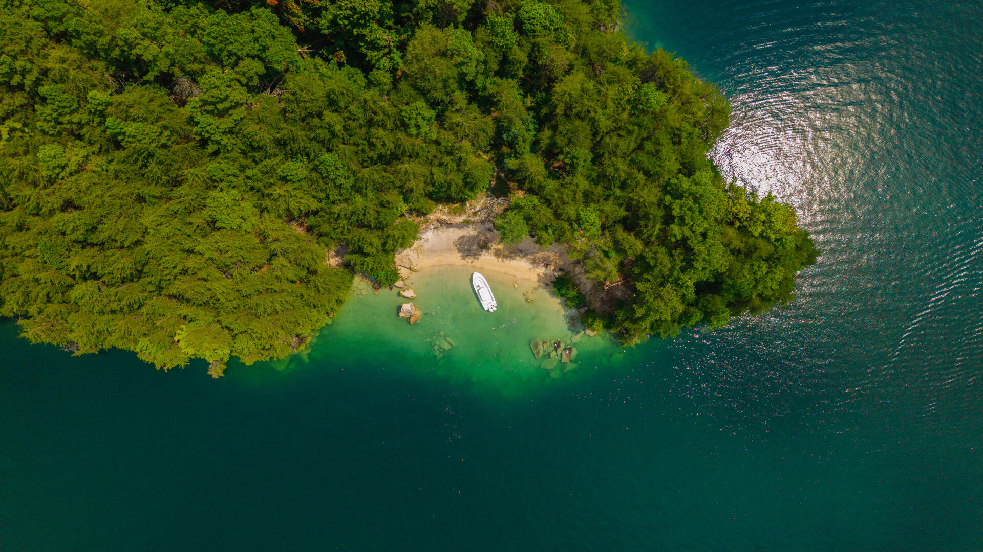 Aerial view of a white boat anchored in turquoise water near a sandy cove surrounded by dense forest