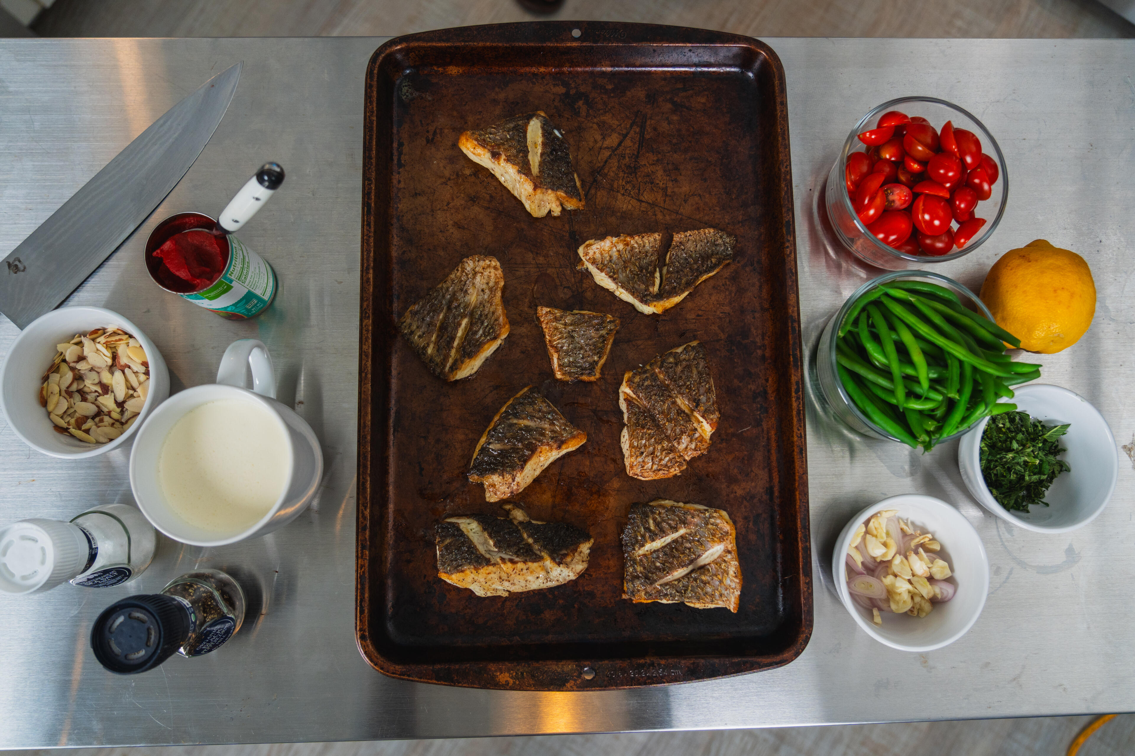 Seared fish fillets on a baking sheet surrounded by ingredients including red peppers, green beans, and almonds on a stainless steel counter
