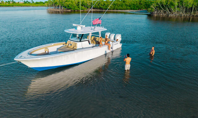 White and blue fishing boat anchored in shallow turquoise water with people swimming nearby and mangroves in the background