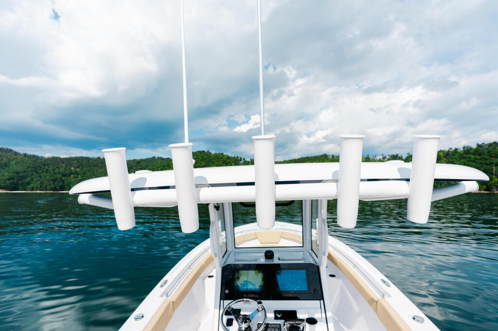 Helm console with dual screens and steering wheel, white hard-top canopy with cylindrical support columns, calm lake waters and tree-lined shoreline beyond