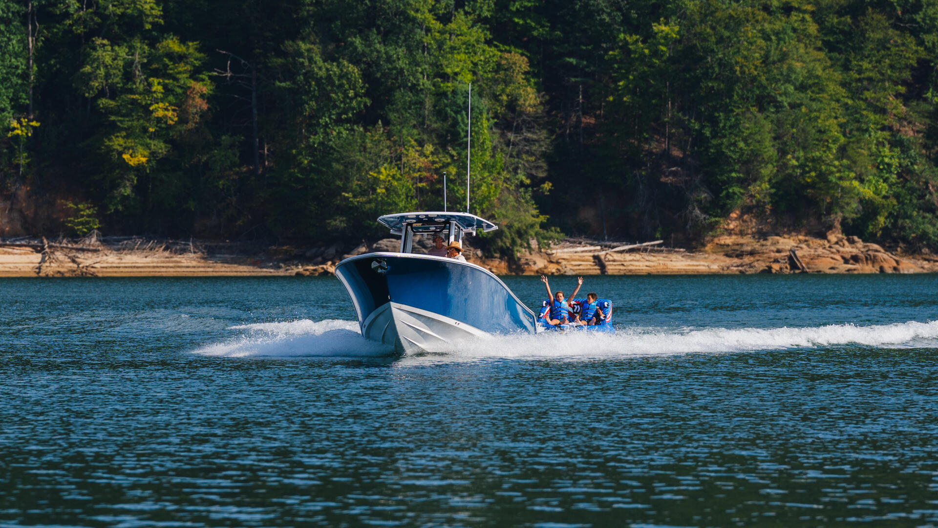 Blue and white speedboat cruising through lake water with passengers waving, forested shoreline in background