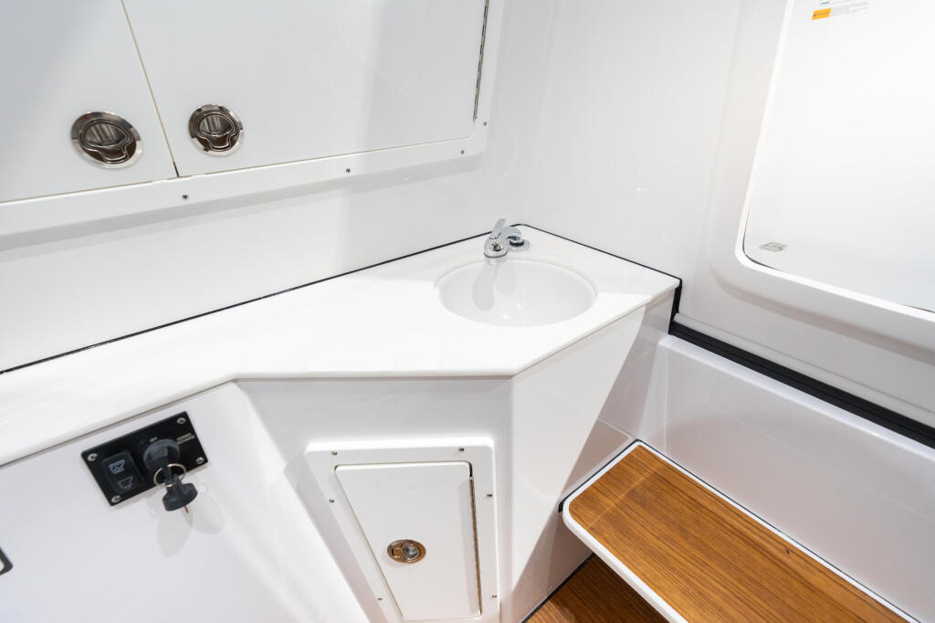 Compact boat cabin with round sink, ventilation vents, control panel, storage hatch, and wood-trimmed seating