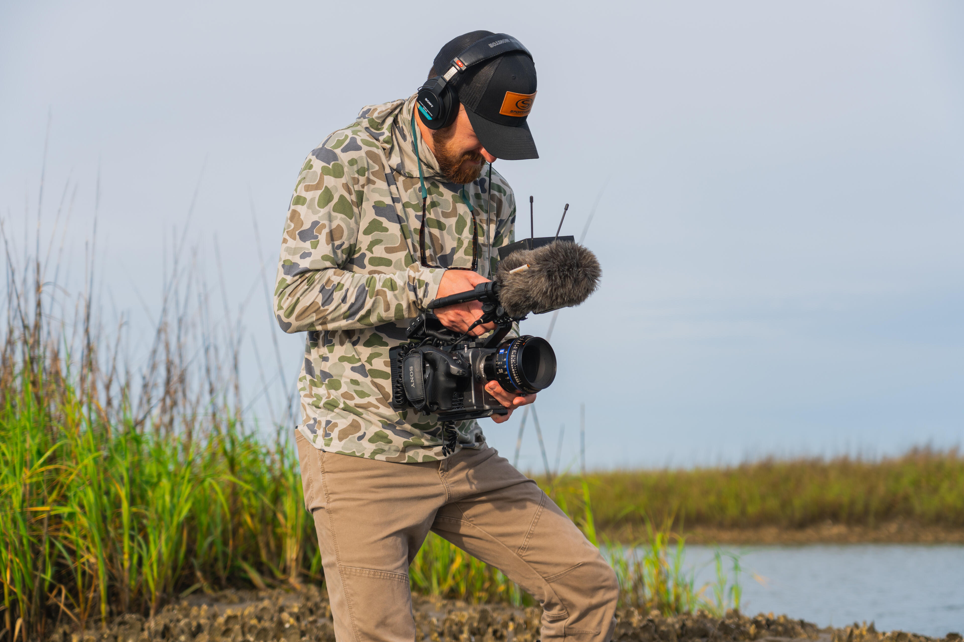Videographer in camouflage jacket records content while standing on a marshy shoreline with camera and wireless audio equipment