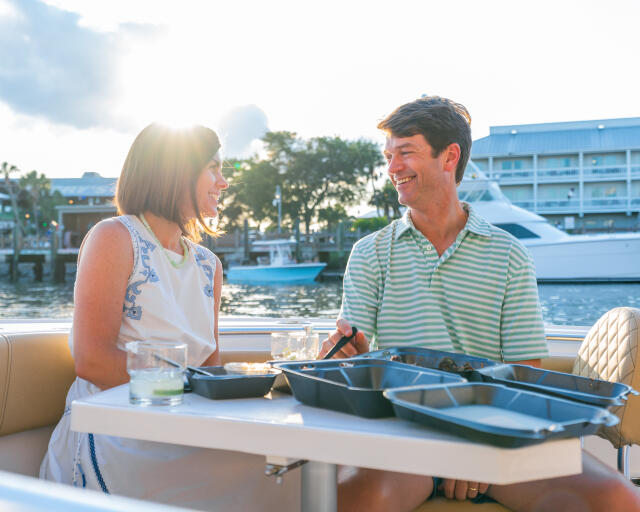 Couple dining at an outdoor table on a boat with marina and waterfront buildings in the background
