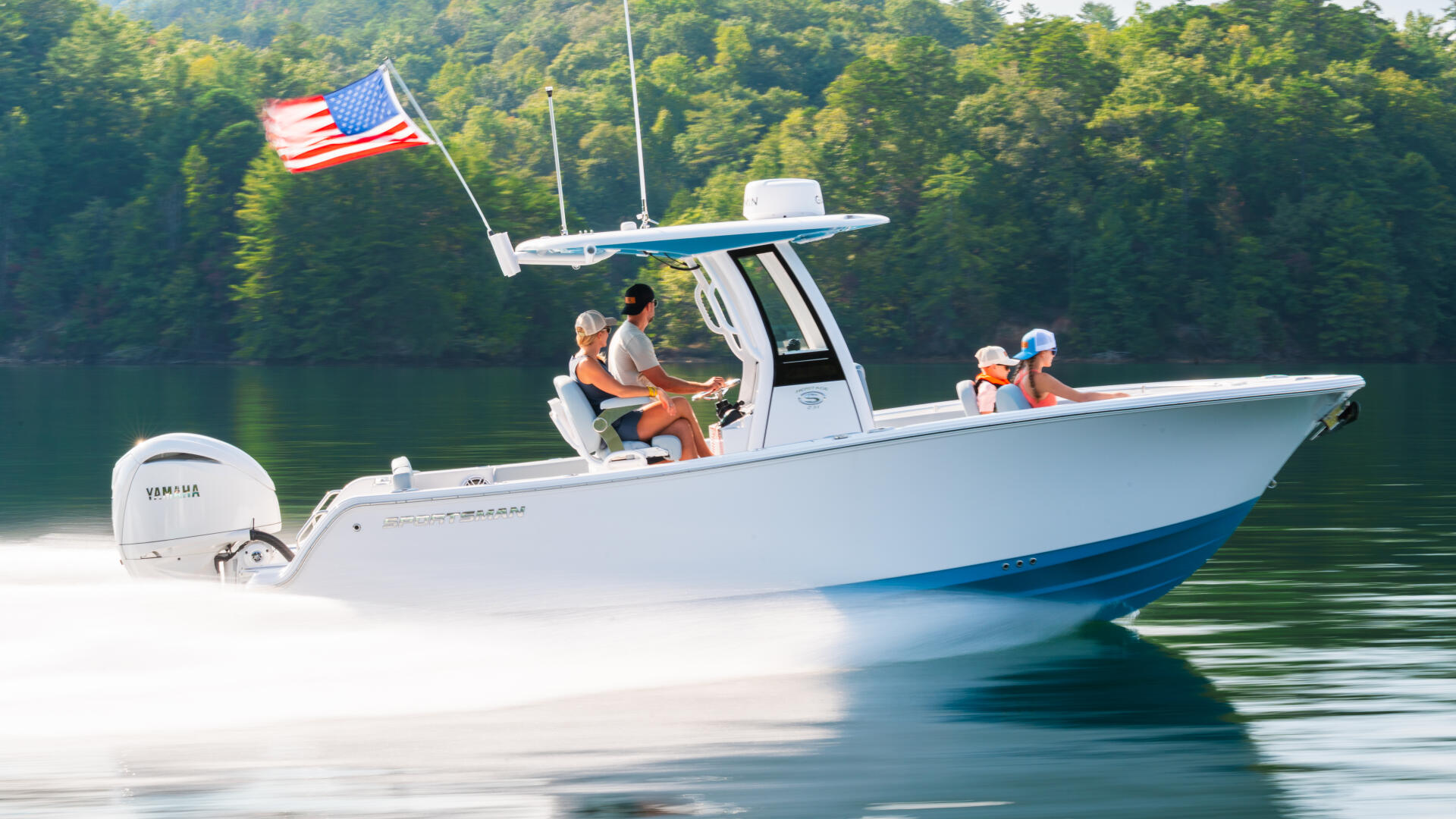 White sportboat with hard-top running at speed on clear lake water, American flag flying, forested mountains in background