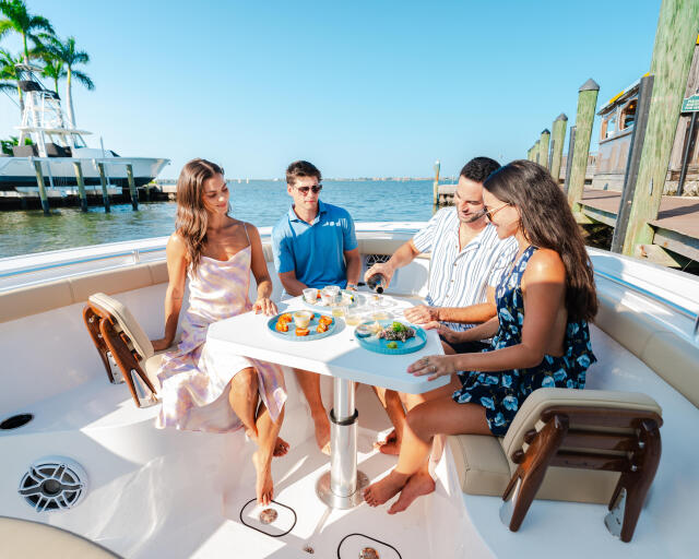 Four friends dining and socializing on a boat docked at a marina with palm trees and waterfront buildings in the background