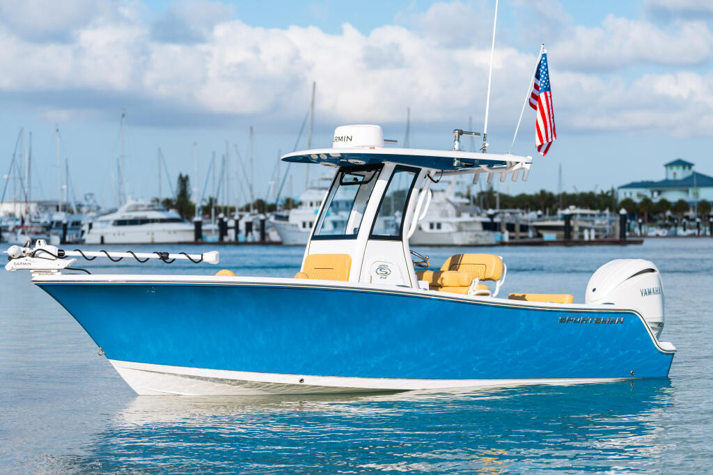 Blue and white center console fishing boat with hard-top anchored in a marina with sailboats in the background