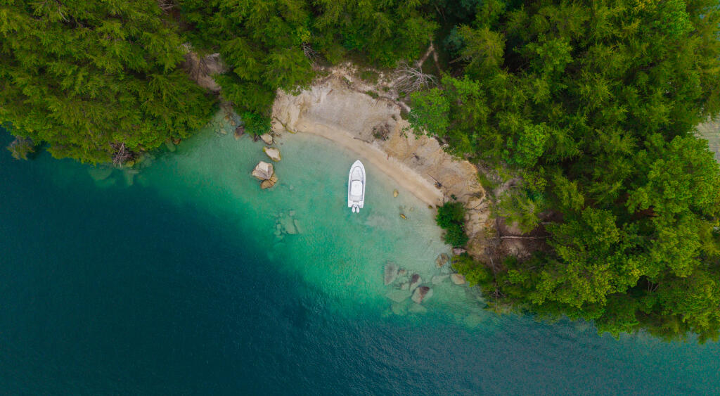 Aerial view of a white boat anchored in turquoise water next to a sandy beach surrounded by dense green forest