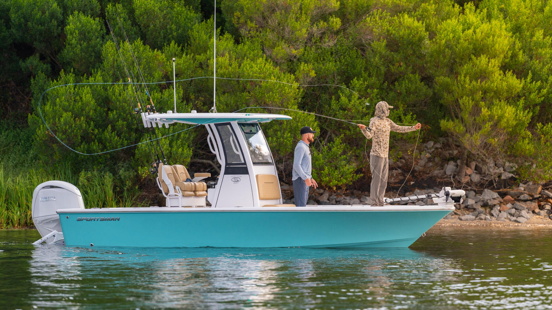 Turquoise boat anchored in shallow water with two anglers fishing from the gunwale near a forested shoreline