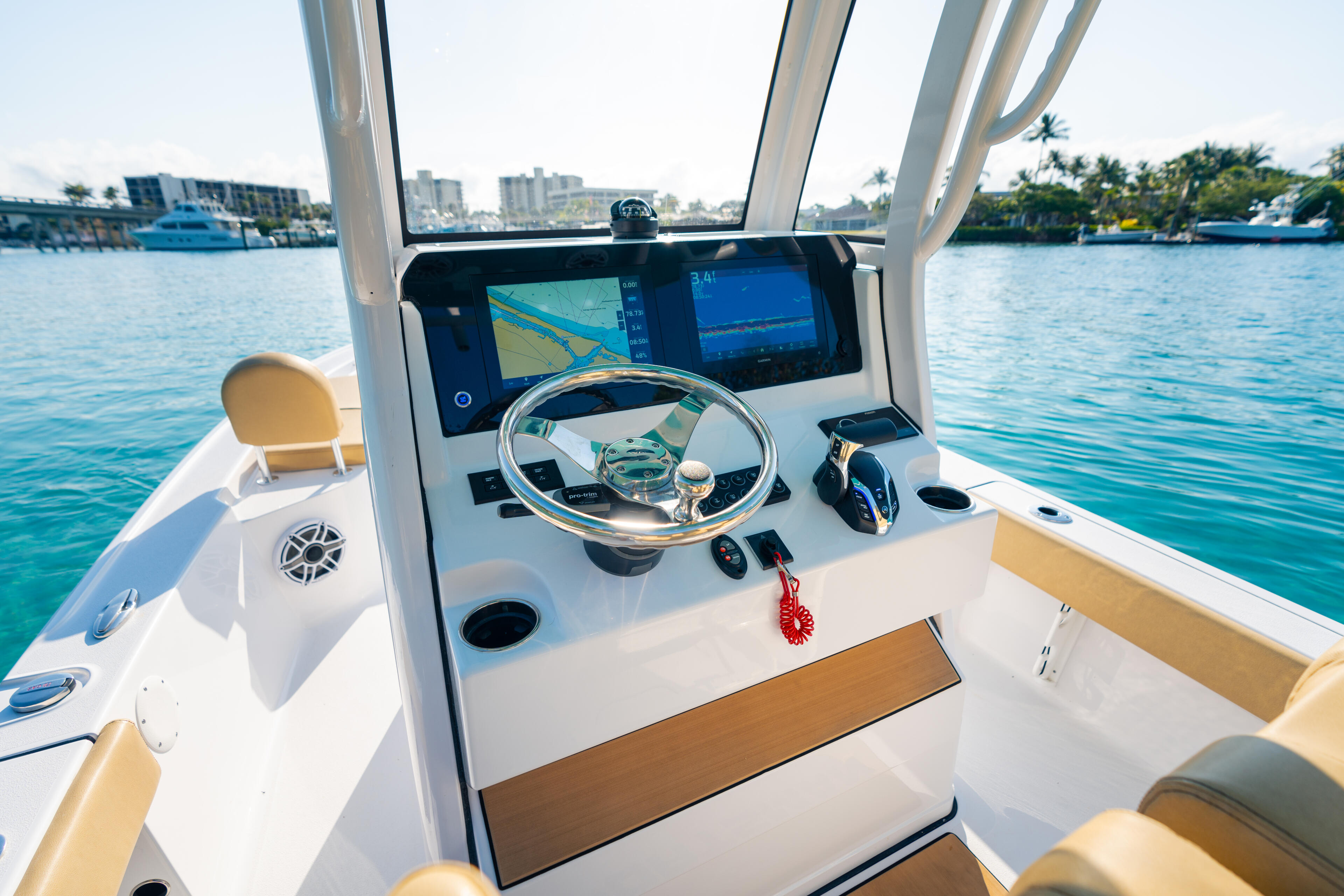 Helm console with dual-screen electronics, steering wheel, and tan seating overlooking a marina with waterfront buildings