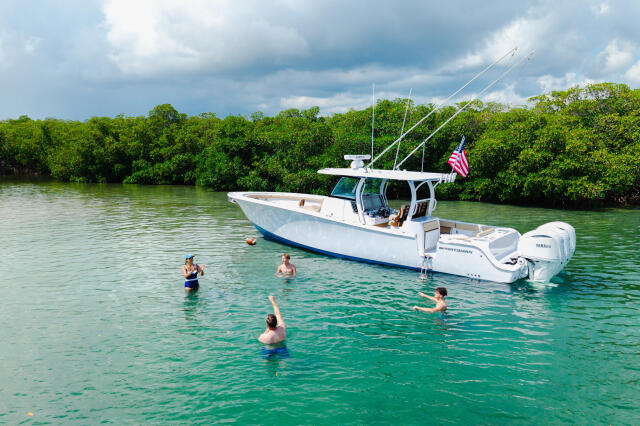 Family swimming and relaxing in turquoise water near a white fishing boat anchored at a sandbar with mangroves in background