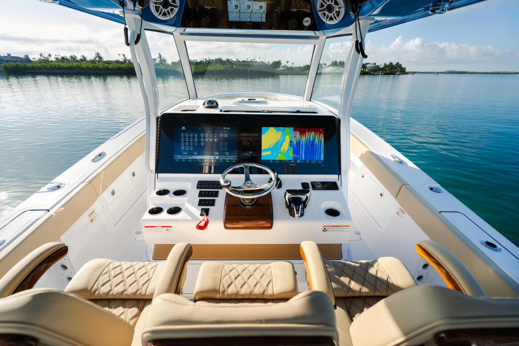Helm of a center console boat with dual monitors, steering wheel, and cream-colored seating facing calm tropical waters