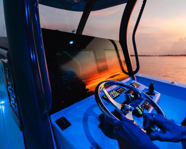 Helm of a blue boat at sunset with illuminated controls and calm waters visible through the windshield