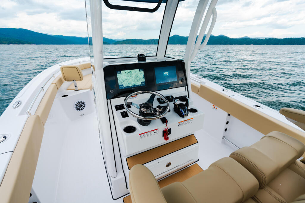 Modern boat helm featuring dual screens, steering wheel, and tan seating with mountain lake and hard-top canopy in background