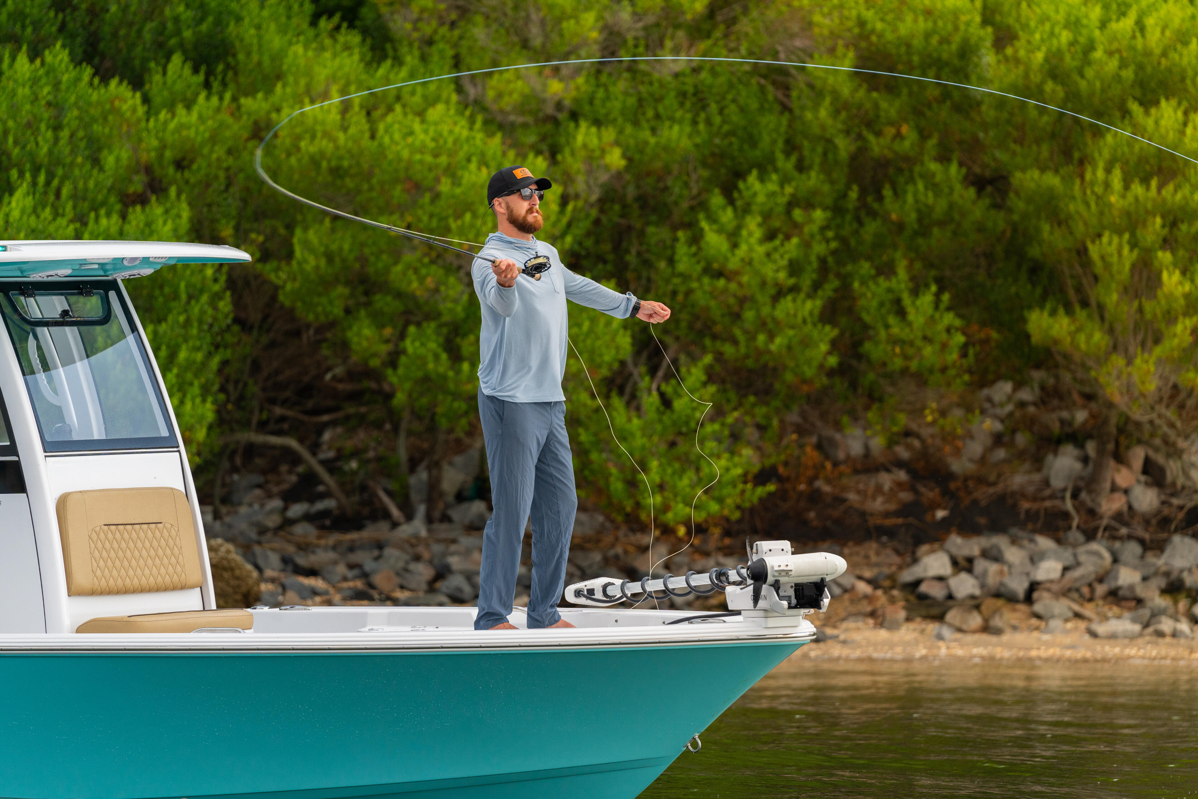 Angler casting a fishing line from the deck of a turquoise and white center console boat near a rocky shoreline