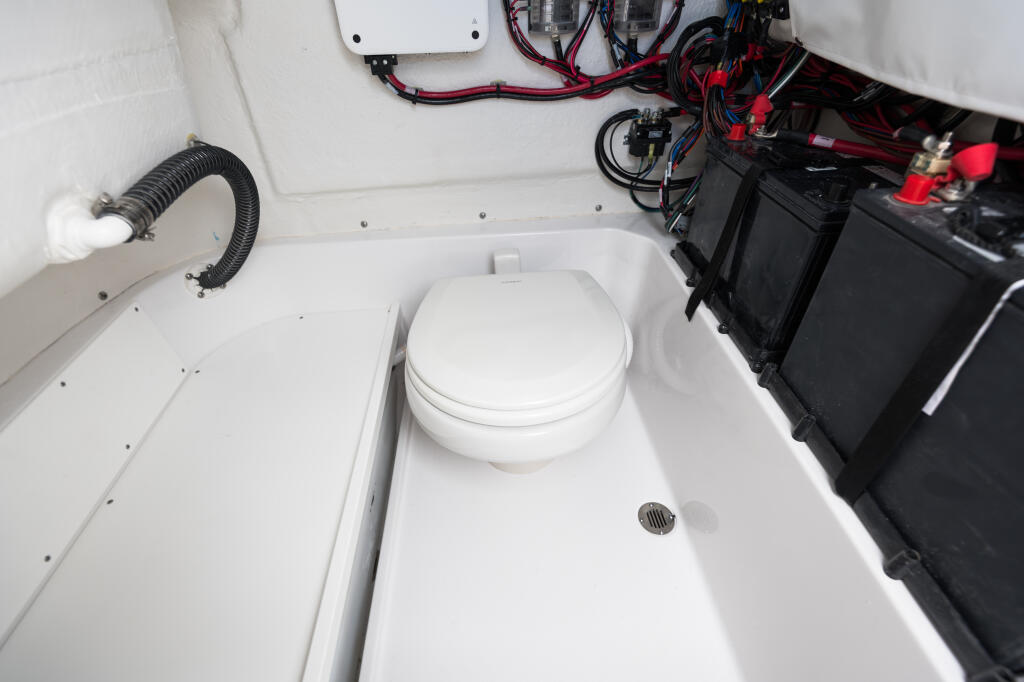 Boat cabin interior with white toilet, battery storage, wiring, and ventilation hose
