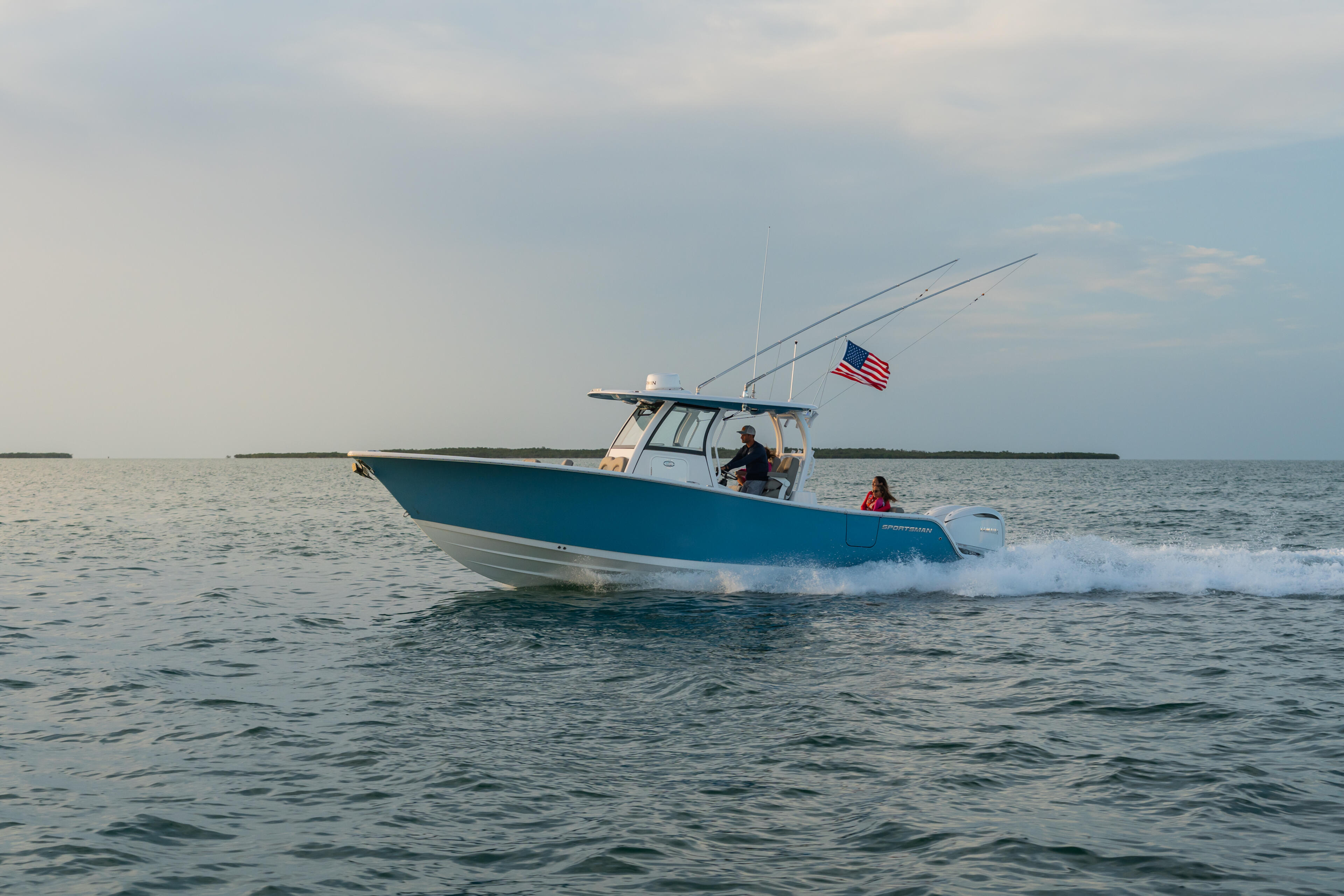 Blue and white fishing boat with hard-top running through shallow water, American flag flying, passengers aboard near sandy shore