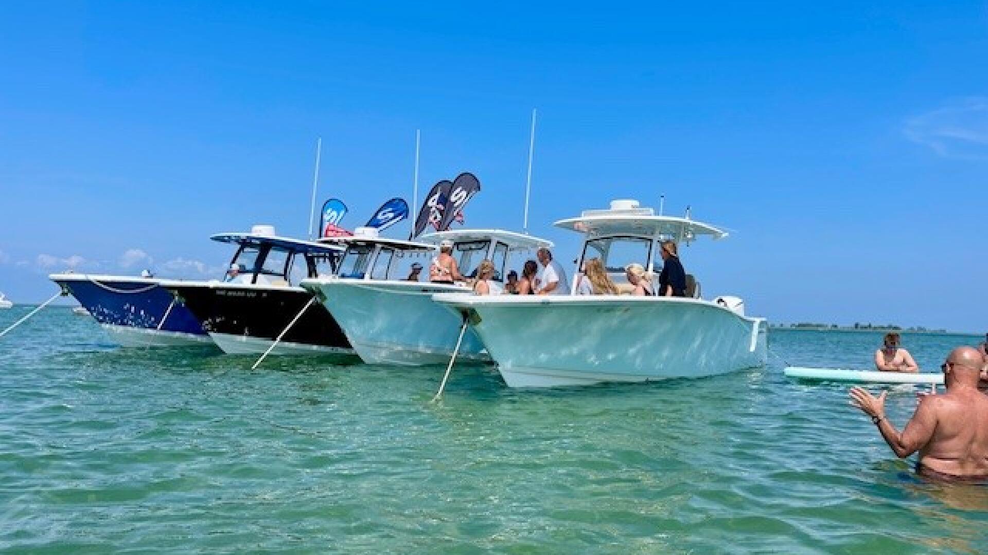 Multiple boats anchored together in shallow turquoise water with people swimming and socializing nearby