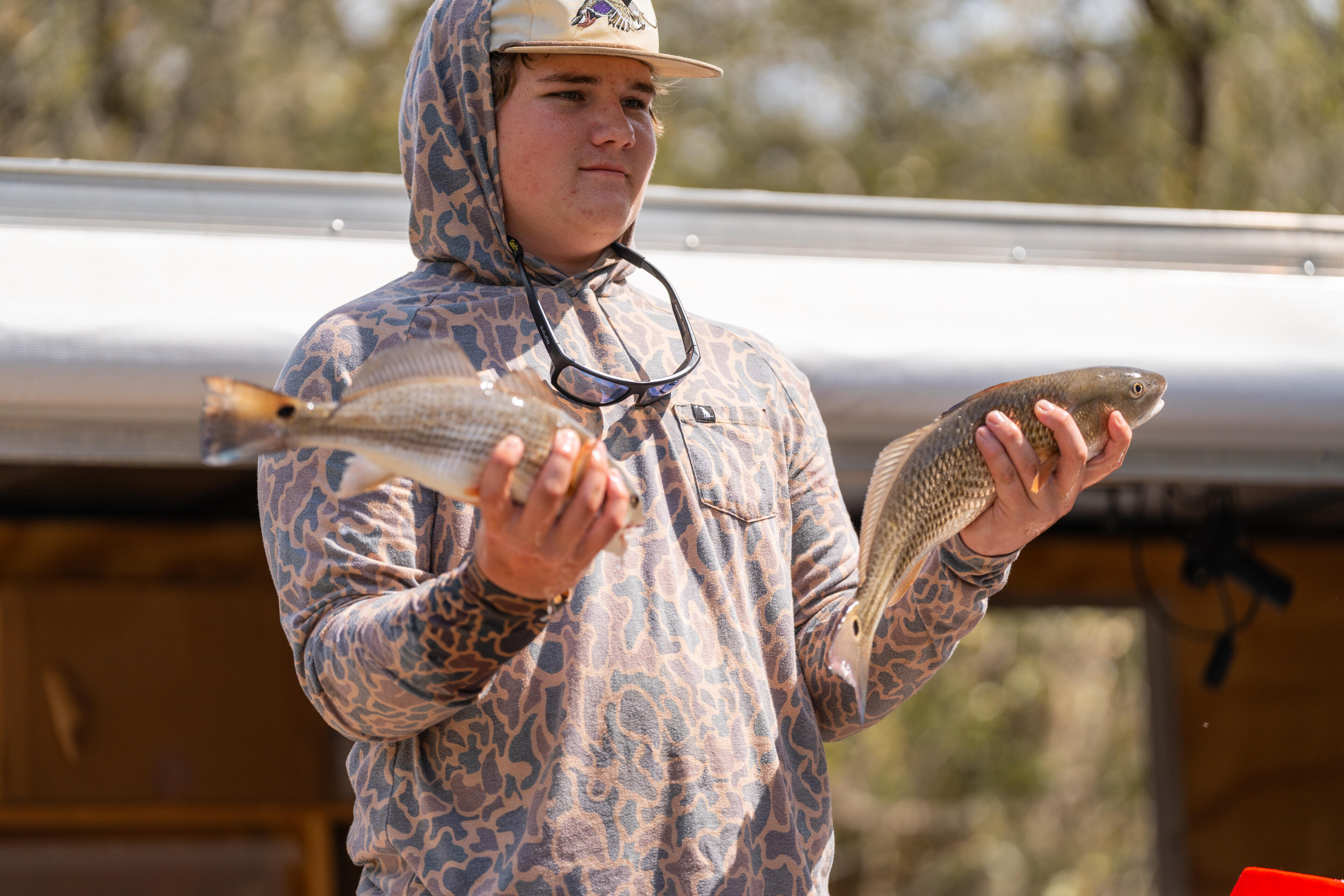 Young angler holding two freshly caught fish on a boat with trees visible in the background