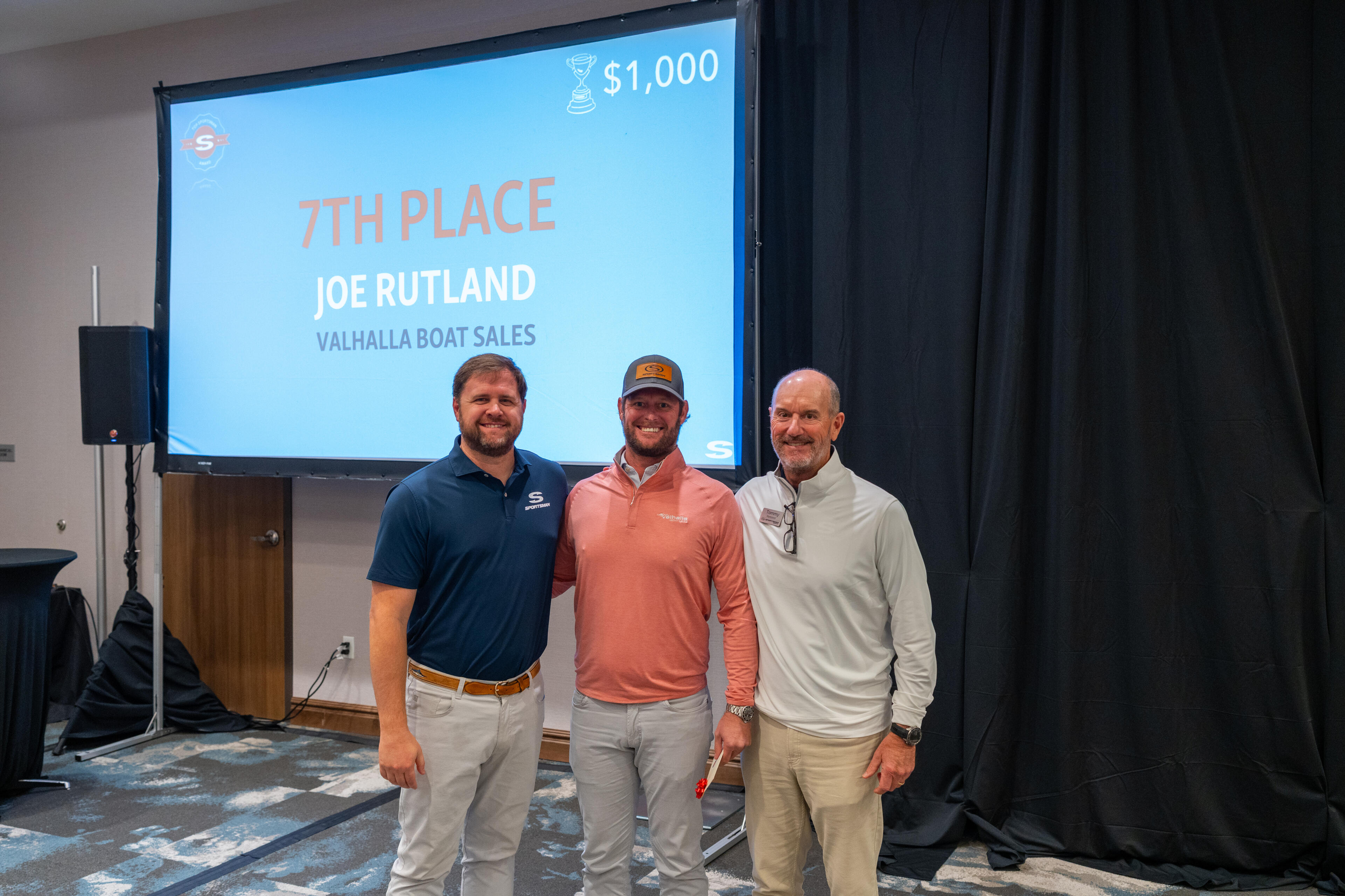 Three men stand in front of a projection screen displaying a 7th Place award for Joe Rutland from Valhalla Boat Sales worth $1,000
