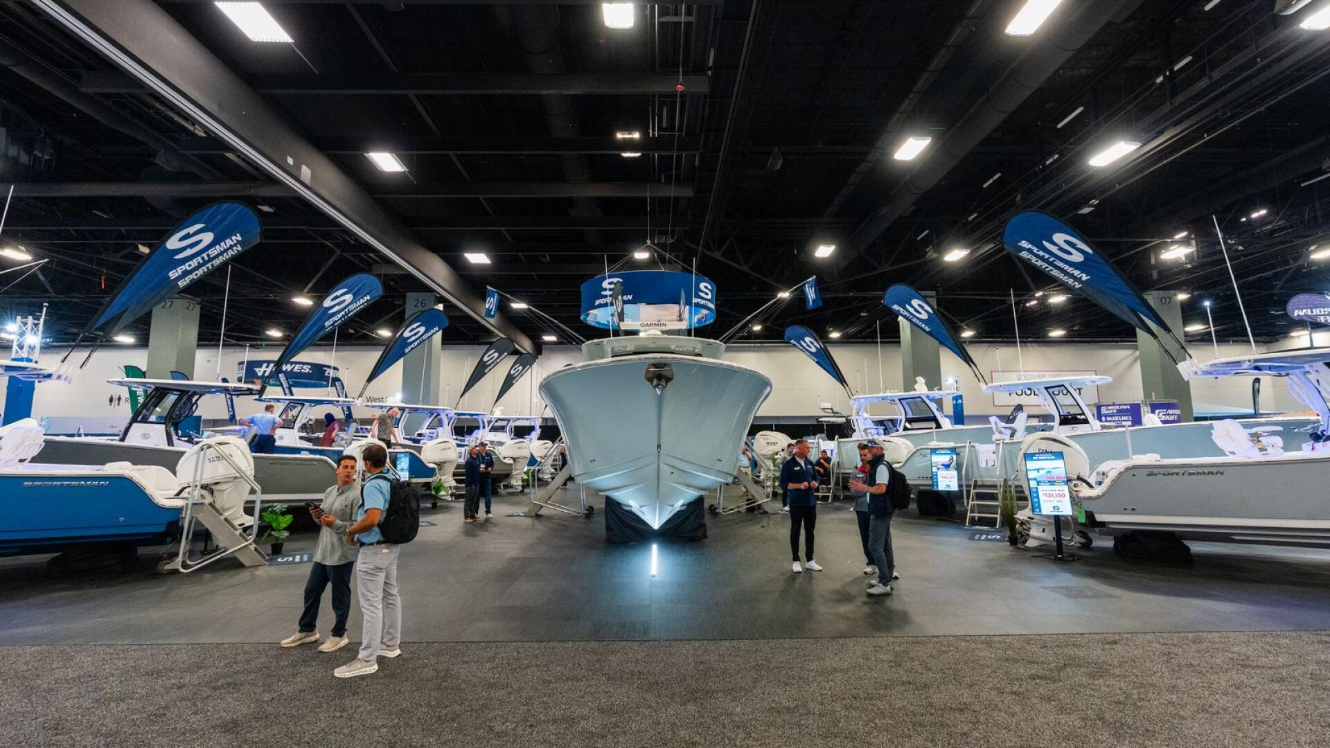Indoor boat show with multiple vessels on display and visitors walking among blue-branded booths and lighting rigs