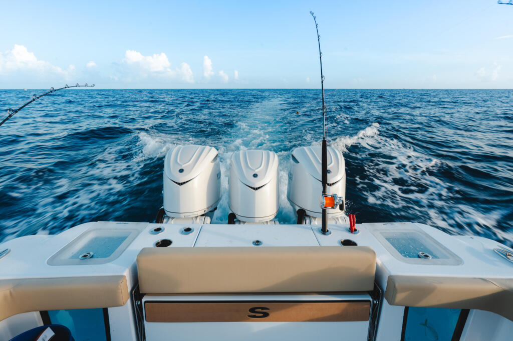 Fishing rod mounted on boat transom with three white engines visible offshore in calm blue waters