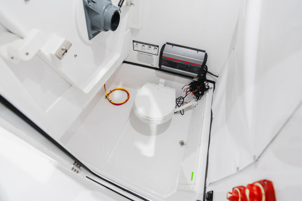 Boat cabin interior with compact toilet, electronics unit, and orange cable against white surfaces