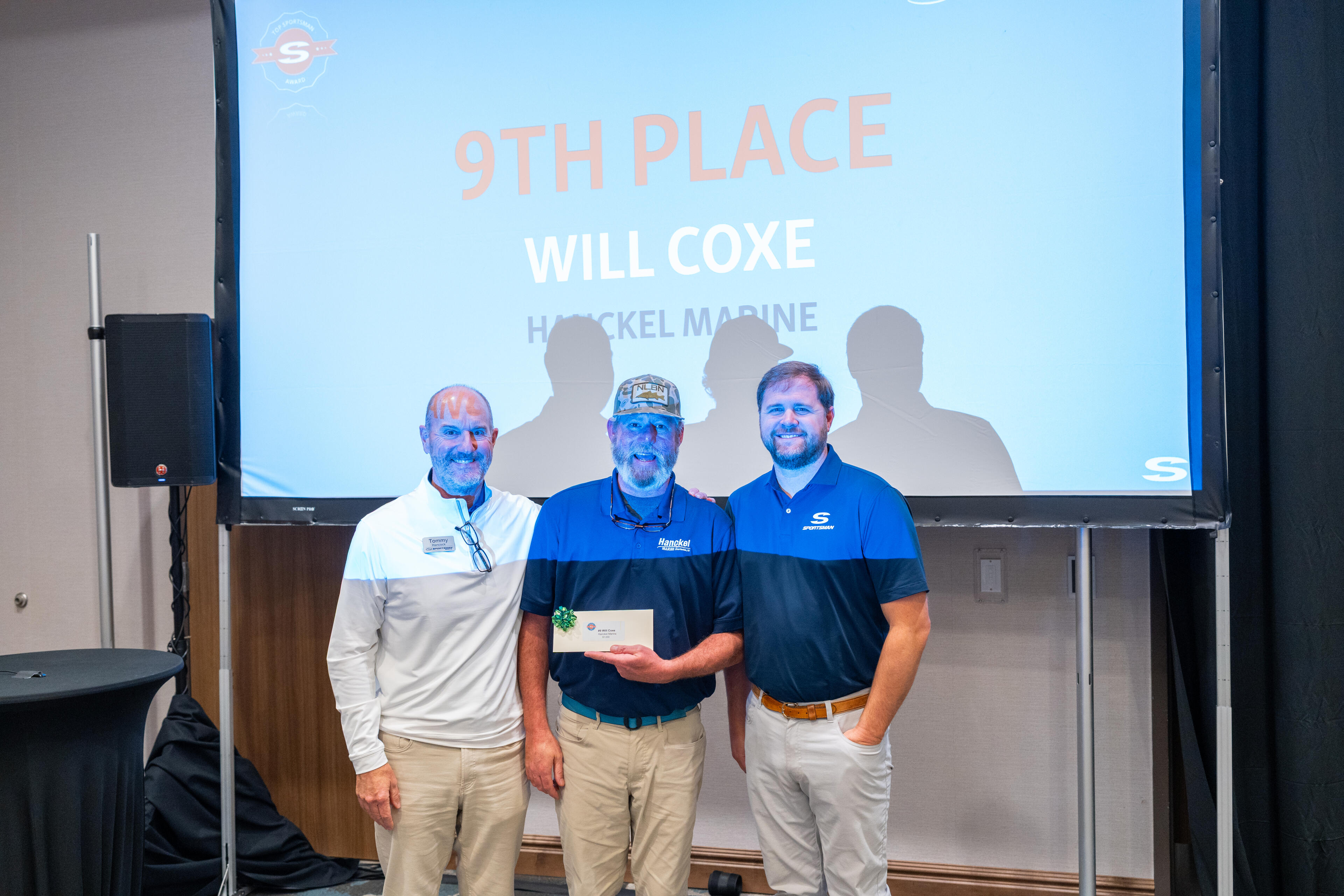 Three men in branded polos stand on stage holding an award with a competition results display projected behind them
