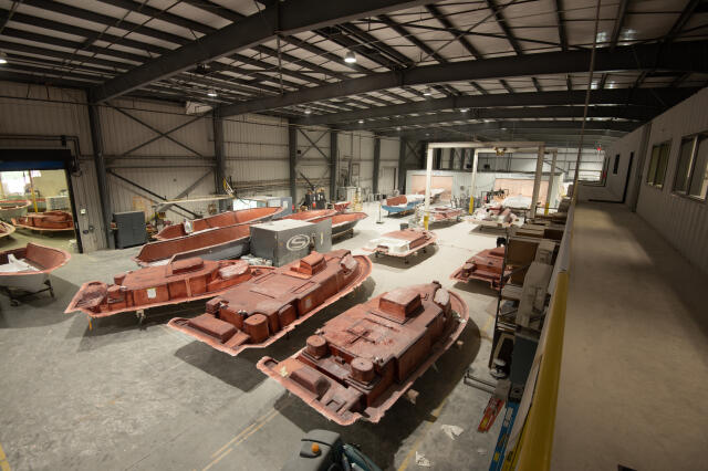 Manufacturing facility with multiple fiberglass boat hull sections in various stages of production under steel structure