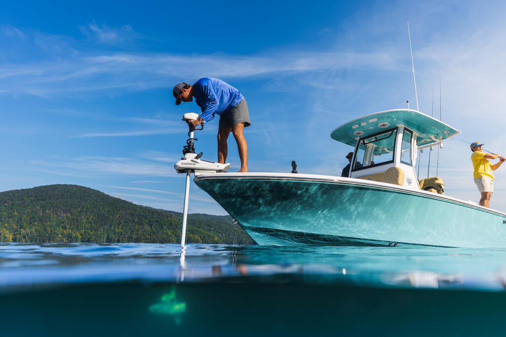 Angler operates a trolling motor from the bow of a turquoise fishing boat on a clear mountain lake