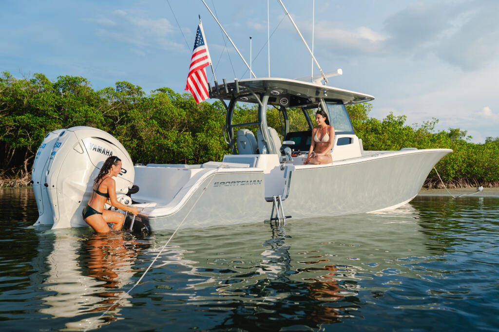 Woman swimming near Sportsman boat with American flag while another person sits at helm near mangroves