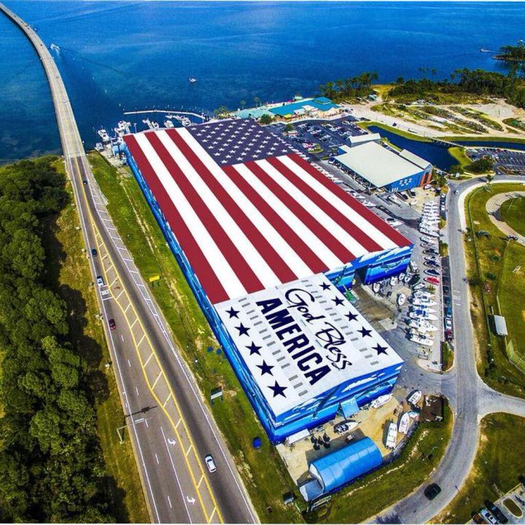 Aerial view of a massive American flag display at a waterfront facility with marina, parking, and coastal infrastructure