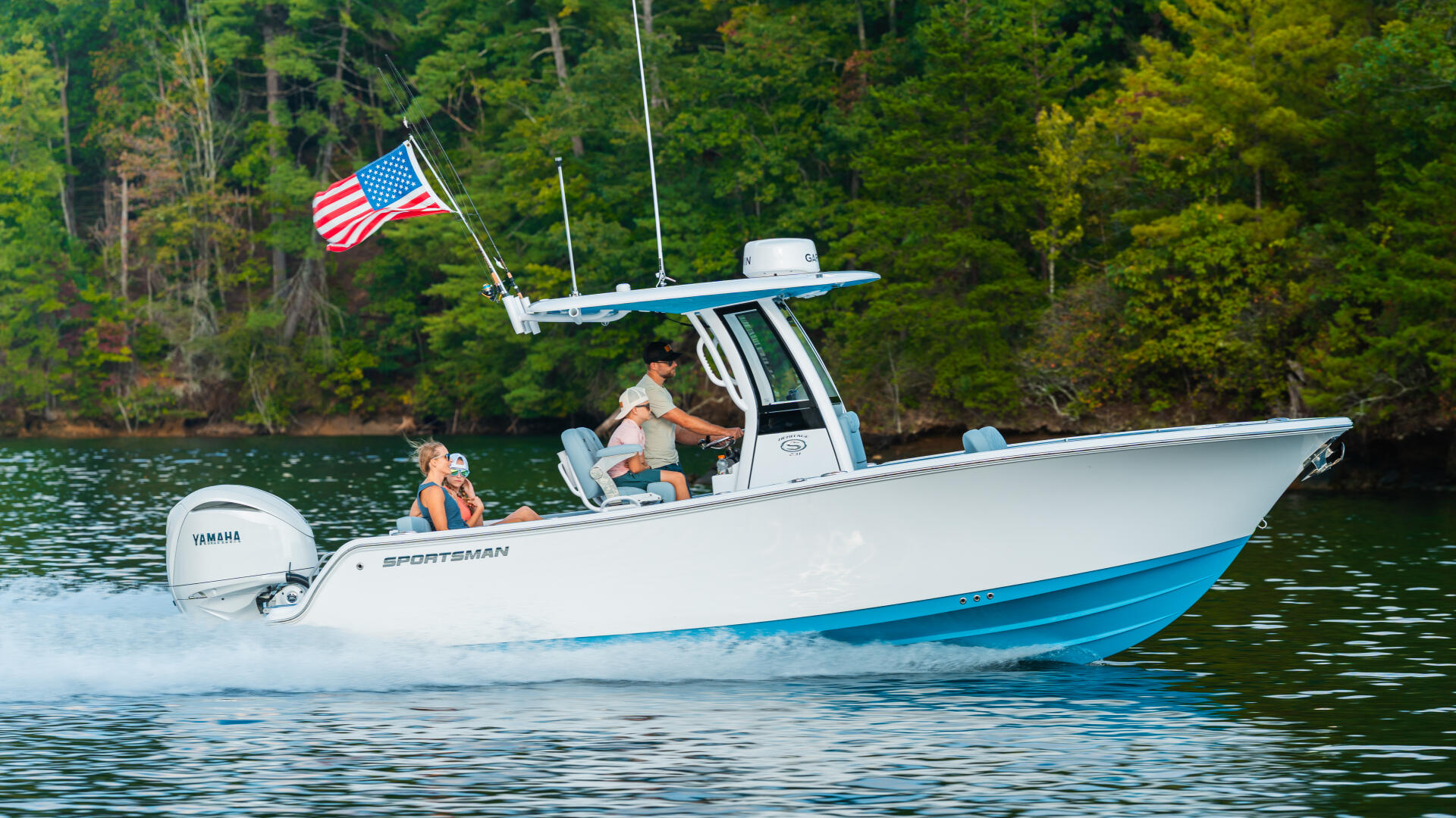 Sportsman boat running on lake with American flag, two people aboard, forested shoreline in background