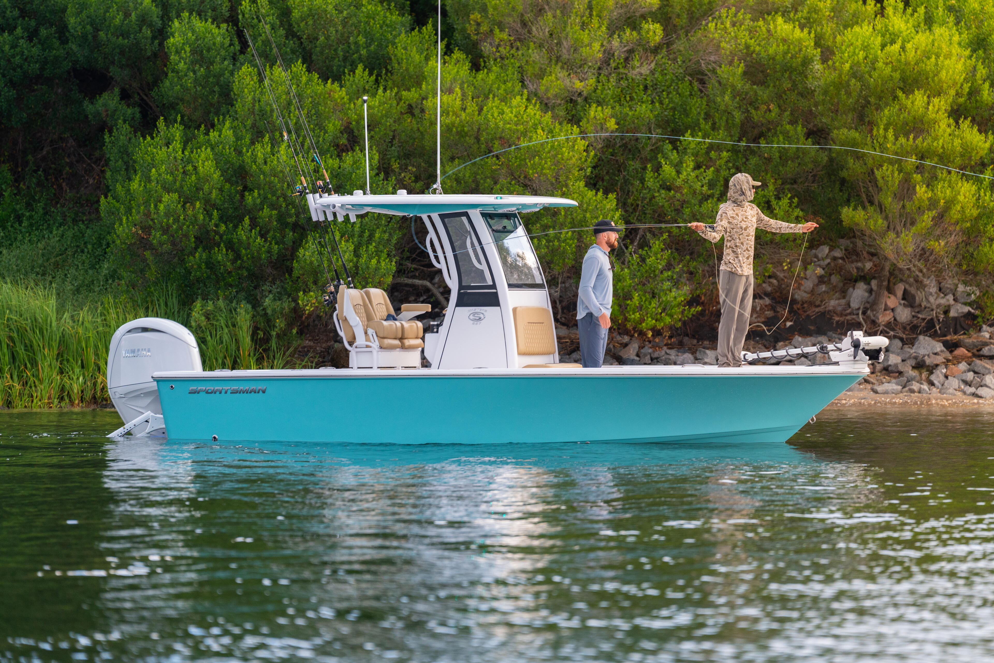 Turquoise fishing boat anchored in shallow water with anglers casting lines near a wooded shoreline
