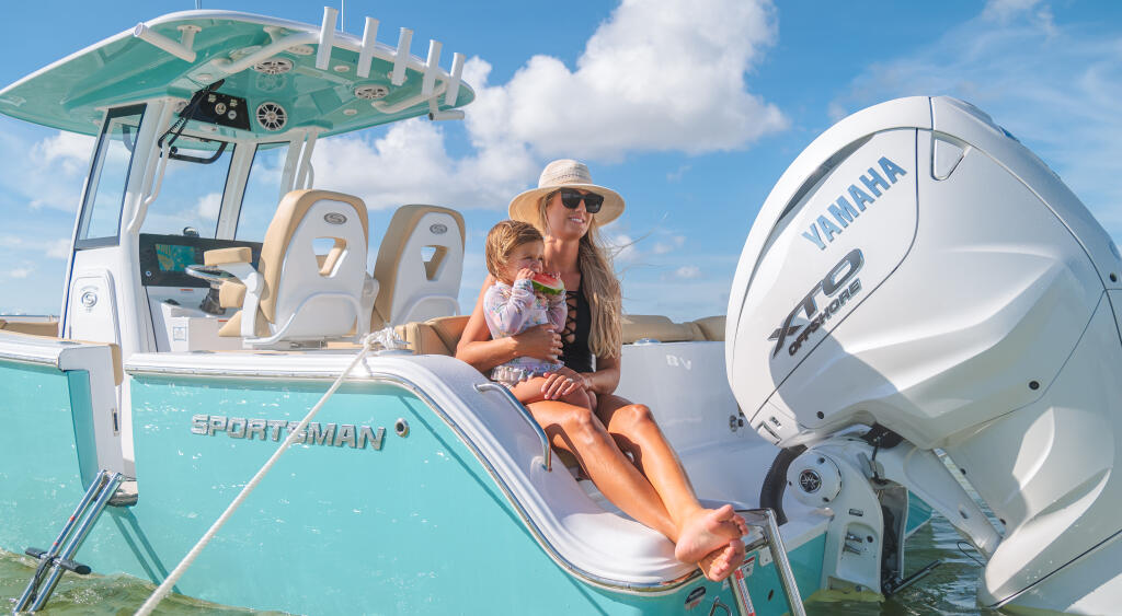 Woman and child sitting on rear seating of turquoise boat at shallow sandbar on clear day