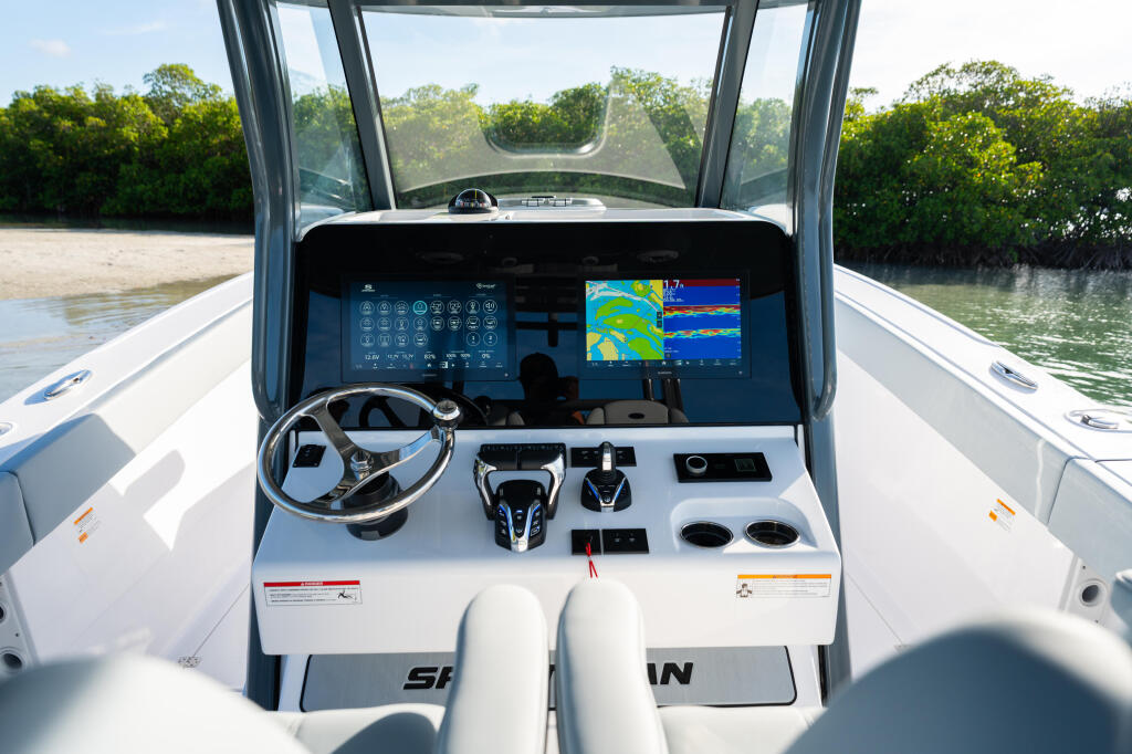 Helm of center console boat with dual monitors showing navigation and sonar displays, steering wheel, and control panel overlooking mangrove shoreline