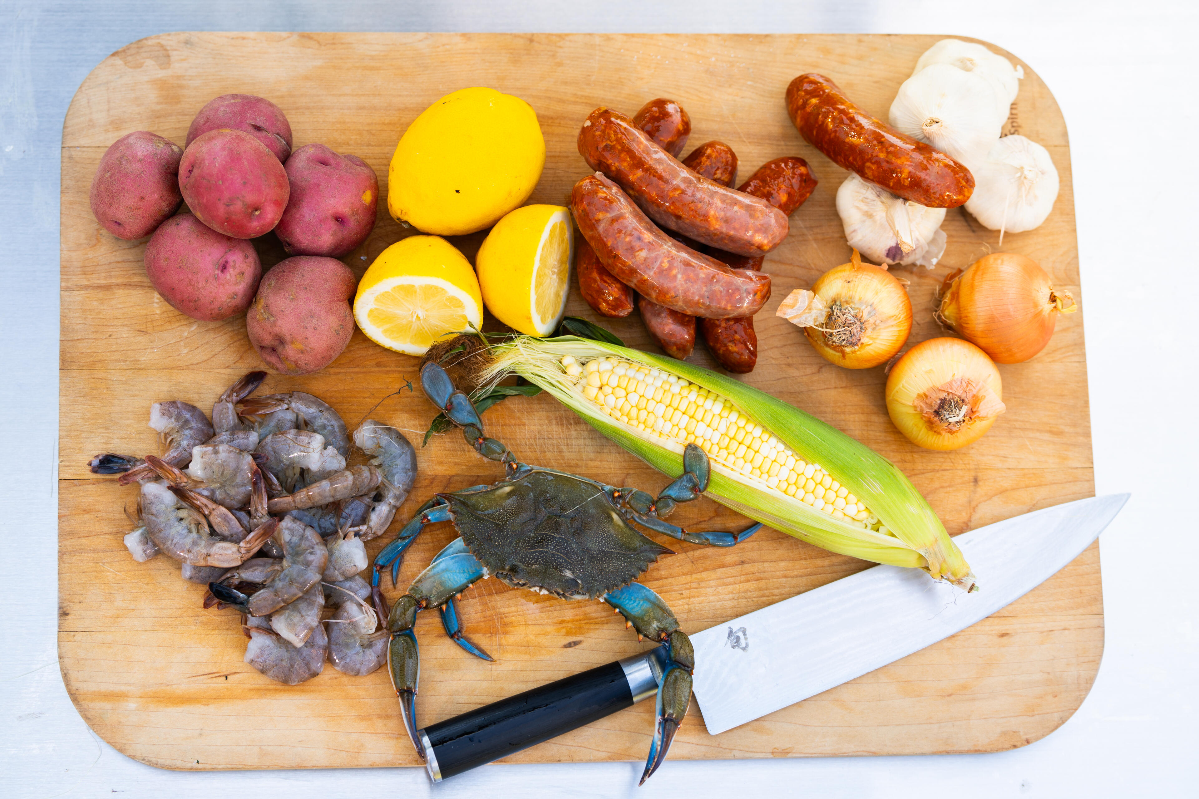 Lowcountry boil ingredients including shrimp, blue crab, corn, sausage, potatoes, mushrooms, and onions arranged on a wooden cutting board