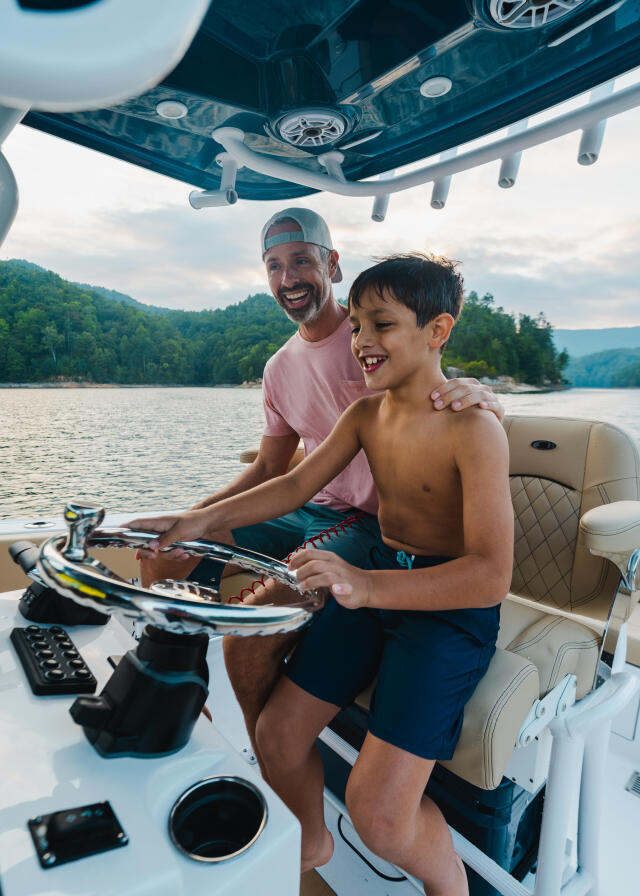 Father and son at the helm of a boat on a lake surrounded by forested mountains, both smiling while holding the steering wheel