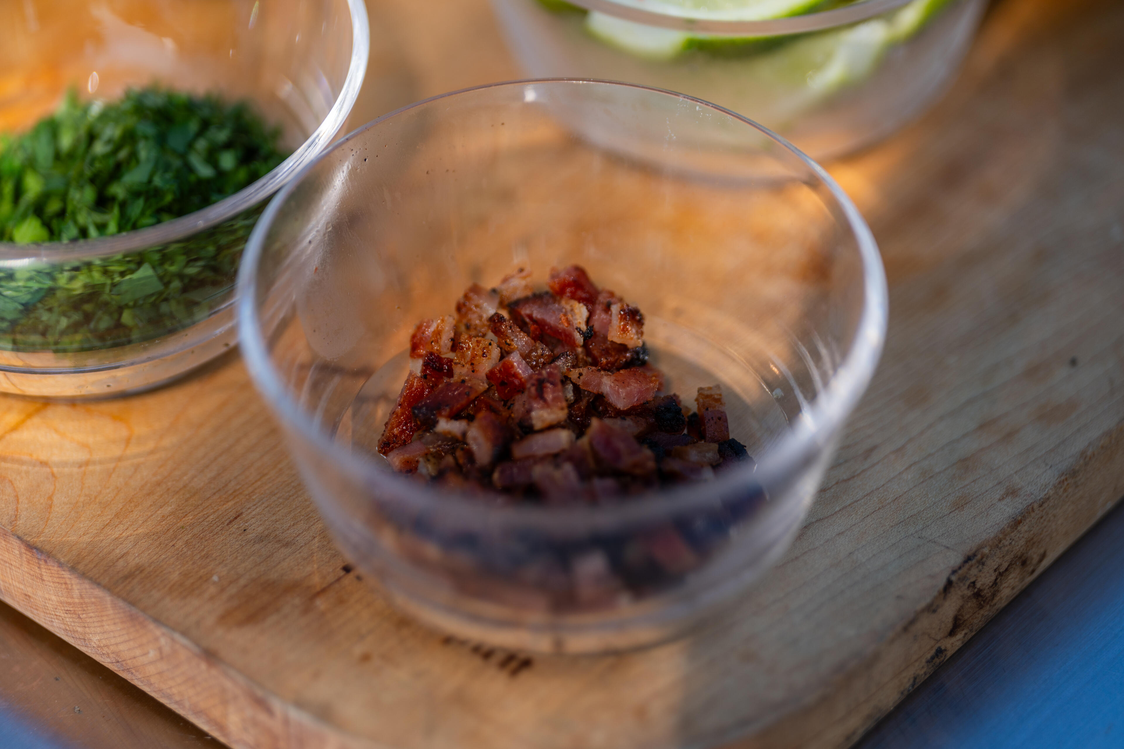 Cooked bacon pieces in a clear bowl with fresh herbs in another bowl on a wooden cutting board